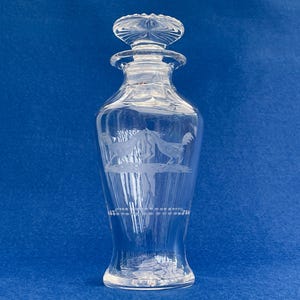 Puede incluir: Una jarra de cristal transparente con tapón, con diseños de gallos grabados. La botella tiene forma bulbosa con cuello estrecho y tapón decorativo. El diseño grabado incluye dos gallos enfrentados. La jarra mide aproximadamente 25 cm de altura.