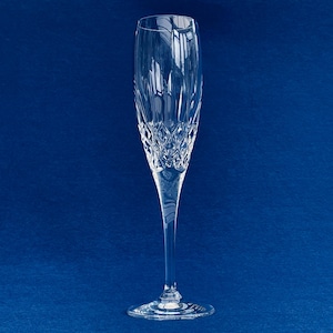 May include: A clear crystal champagne flute with a long stem and a wide, round base. The glass has a diamond-cut pattern on the bowl and vertical lines near the top. The background is a solid, dark blue.