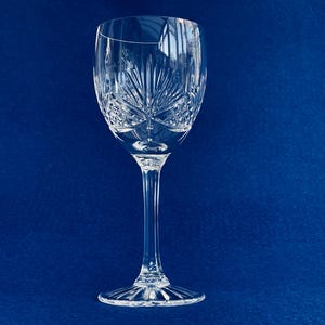 May include: A clear crystal wine glass with a long stem and an etched pattern. The bowl has a starburst design, and the base has a ridged edge. The glass is set against a deep blue background. A classic piece for serving wine.