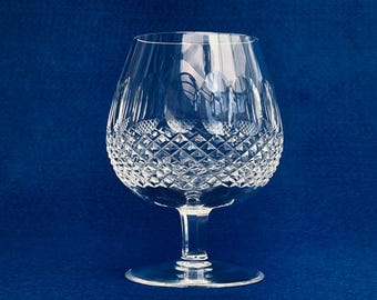 Puede incluir: Una copa de brandy transparente con un patrón de corte de diamante en el cuenco. El vaso tiene un cuenco ancho y redondeado y un tallo corto, sobre un fondo azul oscuro. El vaso está diseñado para realzar el aroma de la bebida.