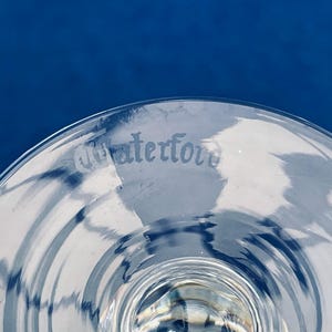 May include: Close-up of a clear glass with the etched word "Waterford" on the rim. The glass has a curved shape and is set against a deep blue background. The glass reflects light, creating a pattern.