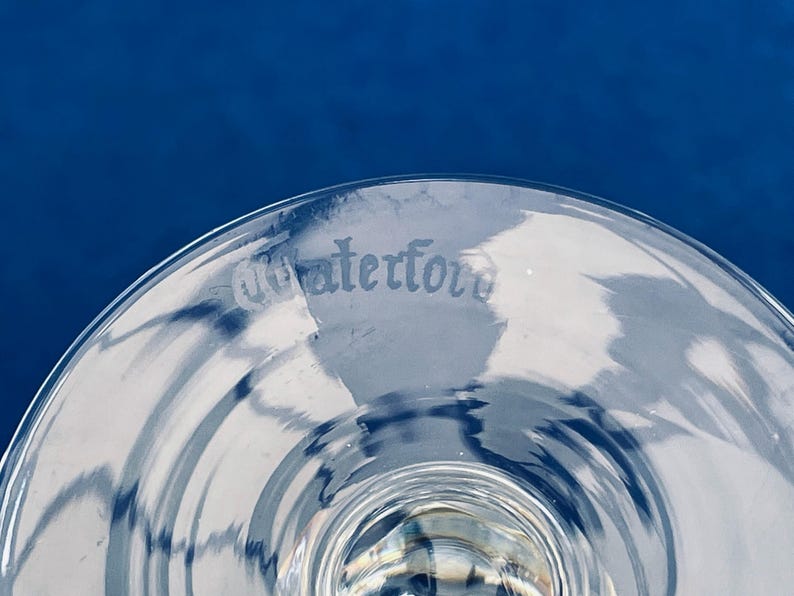 May include: Close-up of a clear glass with the etched word "Waterford" on the rim. The glass has a curved shape and is set against a deep blue background. The glass reflects light, creating a pattern.