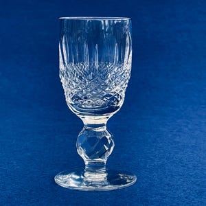 May include: A clear crystal wine glass with a diamond-cut pattern on the bowl and a faceted stem. The glass sits on a round base against a blue background. The glass is approximately 6 inches tall.