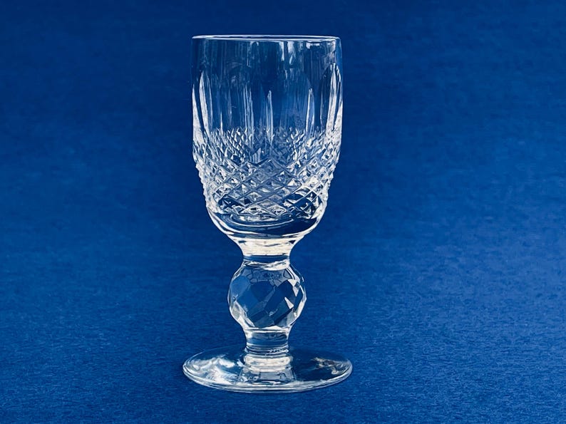 May include: A clear crystal wine glass with a diamond-cut pattern on the bowl and a faceted stem. The glass sits on a round base against a blue background. The glass is approximately 6 inches tall.
