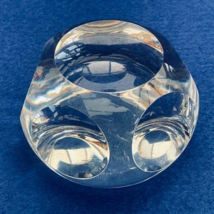 Peut inclure: Une sculpture en verre transparente et géométrique avec des bords arrondis et trois ouvertures circulaires. L'objet transparent reflète la lumière et repose sur une surface bleue. Le design est moderne et abstrait.