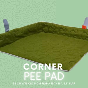 Può includere: Cuscino angolare verde con un motivo trapuntato. Presenta un bordo rialzato e due anelli per appenderlo. Il cuscino misura 38 cm x 38 cm con un lembo di 8 cm. Il testo "CORNER PEE PAD" è visualizzato.
