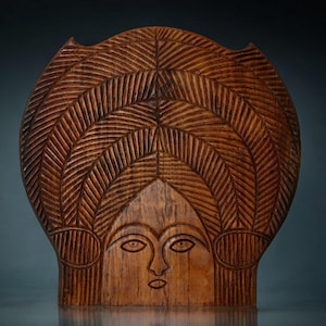 Puede incluir: Una escultura de madera de la cara de una mujer con un peinado tallado detallado. La escultura es de color marrón y tiene un diseño simple y geométrico.