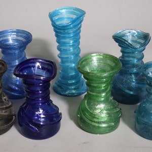 May include: Seven small glass vases in various colors, including blue, green, and gray. The vases have a swirling, textured design and are all different shapes and sizes.