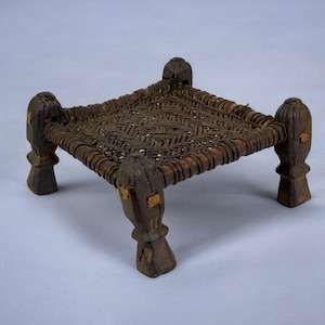 May include: A small, square, woven stool with four legs. The stool is made of dark brown wood and has a woven seat.