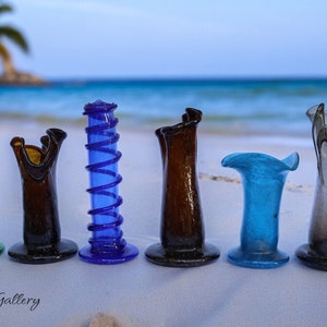 May include: Six small glass vases in various colors and shapes, including green, brown, blue, and gray, arranged in a row on a white sandy beach.