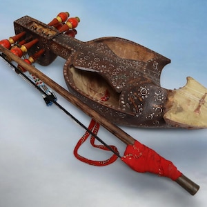 May include: A brown wooden stringed instrument with a carved, ornate design. The instrument has a long, thin neck and a rounded body. It has a red and white string attached to the body. The instrument is a sarangi, a traditional Indian musical instrument.