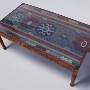 May include: A wooden coffee table with a glass top featuring a colorful patterned rug design. The table has a carved wooden frame and four legs.