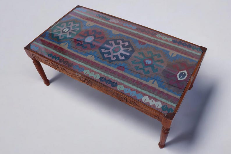 May include: A wooden coffee table with a glass top featuring a colorful patterned rug design. The table has a carved wooden frame and four legs.