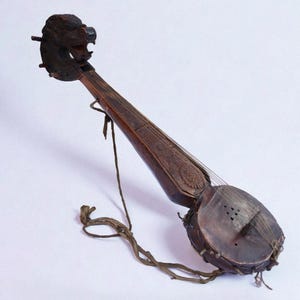 Può includere: Uno strumento a corde antico, marrone scuro, con una testa di leone scolpita in cima. Il corpo è in legno con una cassa armonica circolare e corde. È attaccato un cinturino in pelle.