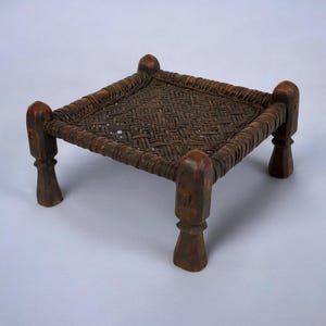 May include: A small, square, wooden stool with a woven seat. The stool has four legs and is made of dark brown wood.