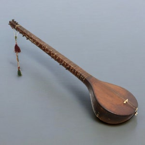 Op de afbeelding: Een langnekkige, houten snaarinstrument met een rond lichaam en een kwast die aan de bovenkant hangt. Het instrument is bruin met een lichtbruine inleg op de hals.