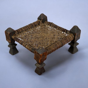 May include: A small, square wooden stool with four legs. The seat is woven with a brown material and the legs are dark brown.