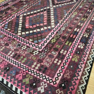 May include: A traditional Turkish rug with a pink, brown, and white geometric pattern. The rug has a fringe border and is made of wool.