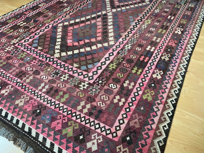 May include: A traditional Turkish rug with a pink, brown, and white geometric pattern. The rug has a fringe border and is made of wool.