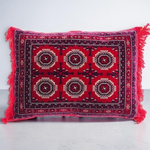 Könnte beinhalten: Ein rechteckiges Zierkissen mit einer leuchtend roten Basis und einem dunkelblauen sowie weißen geometrischen Muster. Das Design umfasst sechs sich wiederholende kreisförmige Motive und eine dekorative Bordüre. Rote Fransen zieren die Seiten.