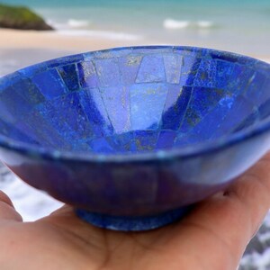 Könnte beinhalten: Eine kleine, blaue Schale aus Lapislazuli, die aus Mosaikfliesen besteht. Die Schale wird in einer Hand gehalten, im Hintergrund sind Strand und Meer zu sehen.