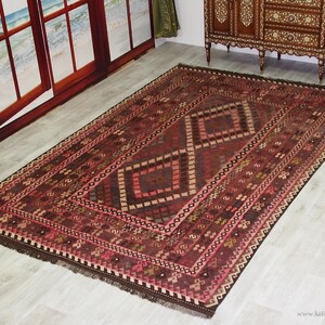 May include: A traditional Turkish rug with a geometric pattern in shades of red, brown, and pink. The rug has a fringe border and is woven with intricate details.