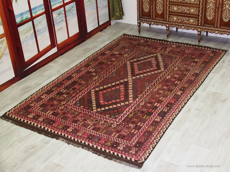 May include: A traditional Turkish rug with a geometric pattern in shades of red, brown, and pink. The rug has a fringe border and is woven with intricate details.