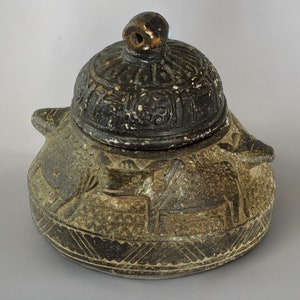 Può includere: Un vaso di ceramica grigia con un coperchio, decorato con figure di animali intagliate e motivi geometrici. Il coperchio ha un piccolo foro nella parte superiore.