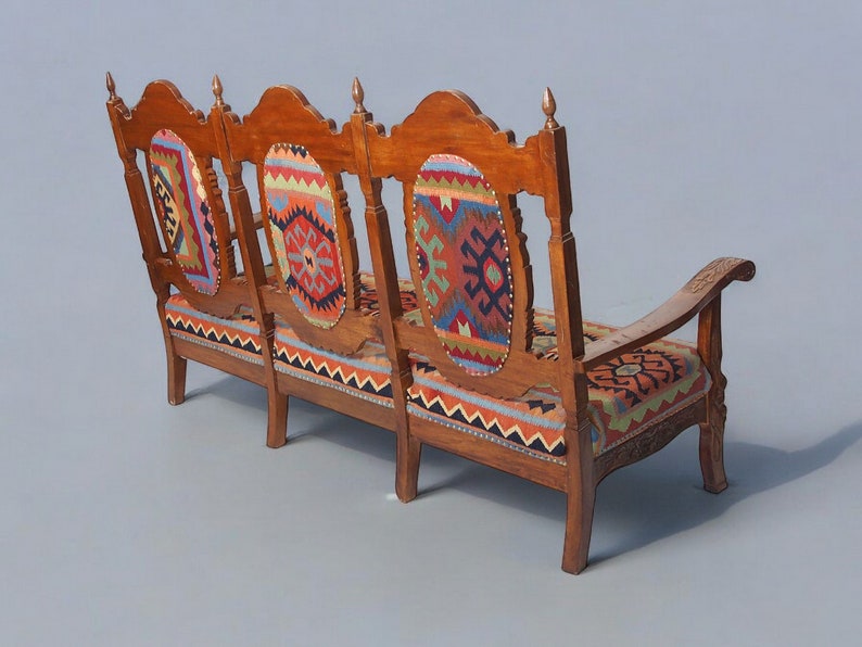 May include: A three-seater wooden sofa with a carved back and armrests. The sofa is upholstered in a colorful patterned fabric with geometric designs in shades of red, blue, green, and yellow.