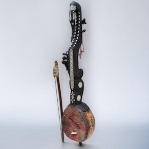 Op de afbeelding: Een handgemaakt snaarinstrument met een lange, sierlijke hals en een afgeronde body. Het instrument is voornamelijk donkerbruin met decoratieve witte accenten. Een strijkstok ligt ernaast. De body heeft een roodbruine tint.