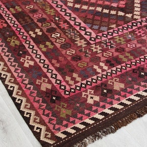 May include: A traditional Turkish rug with a pink, brown, and cream color scheme. The rug has a geometric pattern with intricate details. The rug is hand-woven and has a fringe border.