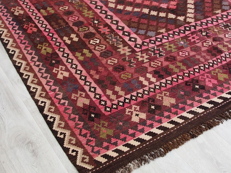 May include: A traditional Turkish rug with a pink, brown, and cream color scheme. The rug has a geometric pattern with intricate details. The rug is hand-woven and has a fringe border.