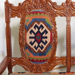 May include: A wooden chair with intricate carvings and a colorful patterned cushion. The chair has a high back and a wide seat. The cushion is made of a woven fabric with geometric designs in shades of red, blue, green, and brown.