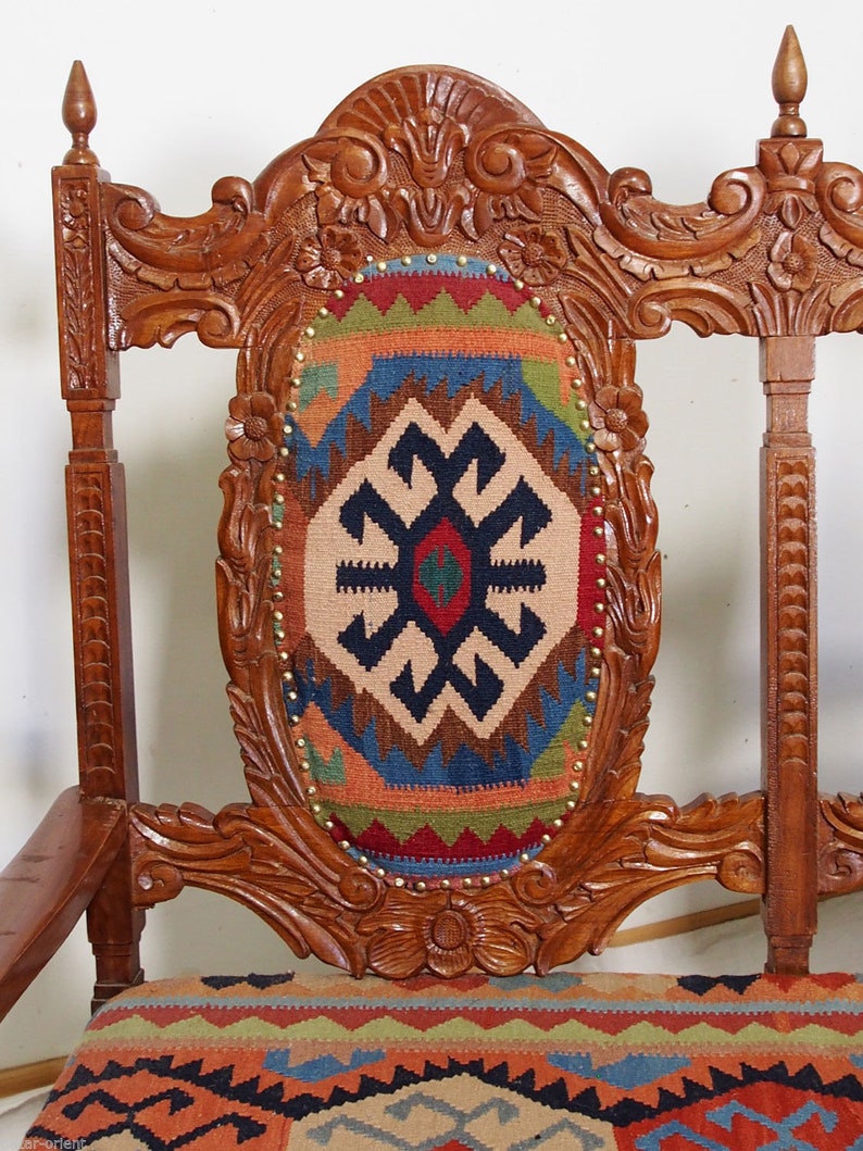 May include: A wooden chair with intricate carvings and a colorful patterned cushion. The chair has a high back and a wide seat. The cushion is made of a woven fabric with geometric designs in shades of red, blue, green, and brown.
