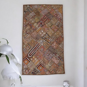 Puede incluir: Un tapiz rectangular con un diseño de patchwork. El tapiz presenta una variedad de patrones geométricos en tonos marrones, rojos, naranjas y dorados. Se incorporan pequeños espejos en el diseño, agregando un elemento reflectante.