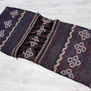 Op de afbeelding: Een donkerblauw en bruin geweven tapijt met een geometrisch patroon en franjes. Het tapijt heeft een traditioneel ontwerp met rode, witte en crème accenten.