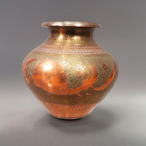 May include: A copper and brass vase with intricate etched designs. The vase has a rounded body and a narrow neck. The designs are in a floral pattern and include a peacock.