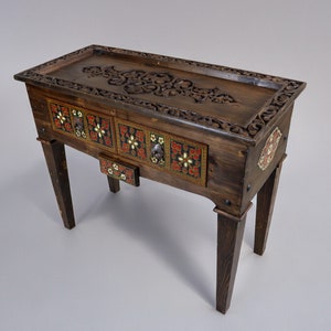 Puede incluir: Una mesa de madera con tres cajones que presentan intrincados diseños de azulejos florales en rojo, negro y dorado. La mesa tiene una tapa de madera tallada y cuatro patas.