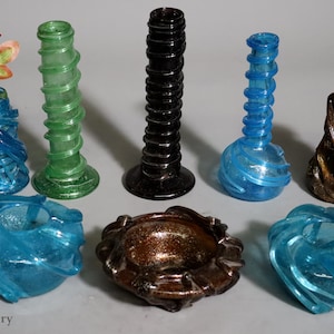 May include: A collection of seven glass vases in various colors and designs. The vases are all made of clear glass with a spiral pattern. The vases are arranged in a circle on a white surface. Kabul Gallery is printed at the bottom of the image.
