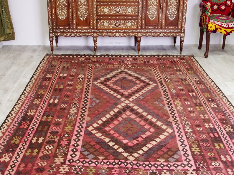 May include: A traditional red, pink, and brown patterned rug with a diamond-shaped design in the center. The rug has a fringe border.