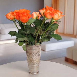 Puede incluir: Un ramo de rosas naranjas en un jarrón plateado con un diseño ornamentado. El jarrón está sobre una mesa blanca.