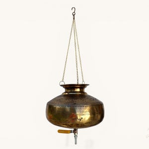 Könnte beinhalten: Eine hängende Wasserkanne aus Messing mit Zapfhahn und dekorativem, geätztem Design. Die Kanne wird von einer Kette an einem Haken aufgehängt. Der Zapfhahn hat einen gelben Griff.