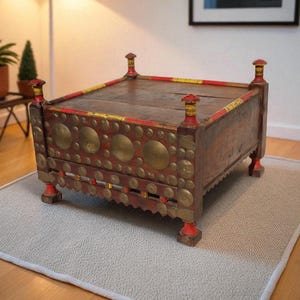 May include: An antique wooden chest with red, gold, and brass accents. The chest has a rectangular shape with decorative brass circles and is placed on a light gray rug. The chest has four decorative posts on each corner.
