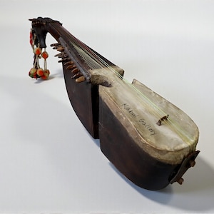 Op de afbeelding: Een snaarinstrument met een houten body en hals, een lichtgekleurde klankkast en donkerbruine zijkanten. Het instrument heeft meerdere snaren en een decoratieve kop met hangende kralen. De tekst "Kabul Gallery" is zichtbaar.