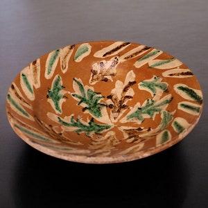 Può includere: Una ciotola in ceramica marrone con un motivo floreale verde e marrone. La ciotola ha un bordo leggermente rialzato e una superficie liscia e smaltata.
