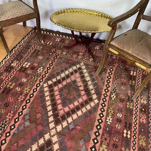 May include: A vintage rug with a geometric pattern in shades of pink, brown, and black. The rug is laid out on a wooden floor with two antique chairs and a gold tray table.