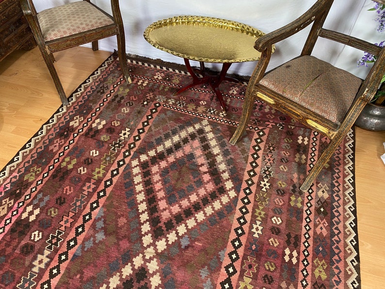 May include: A vintage rug with a geometric pattern in shades of pink, brown, and black. The rug is laid out on a wooden floor with two antique chairs and a gold tray table.