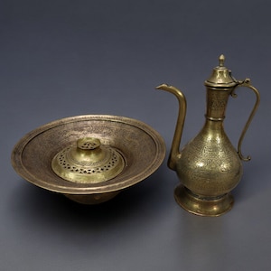 Può includere: Un bruciatore di incenso in ottone con un coperchio decorativo e una brocca in ottone abbinata con un design ornamentale. Entrambi i pezzi sono incisi con motivi intricati.