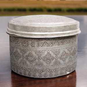 May include: A silver-colored, round container with a lid. The container's body is adorned with an intricate, repeating floral and geometric pattern. The lid and body are made of metal. The container is likely used for storage.