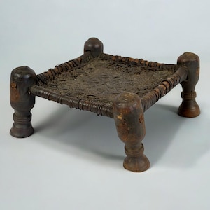 May include: A small, square wooden stool with four turned legs and a woven seat. The stool is made of dark brown wood and has a rustic appearance.